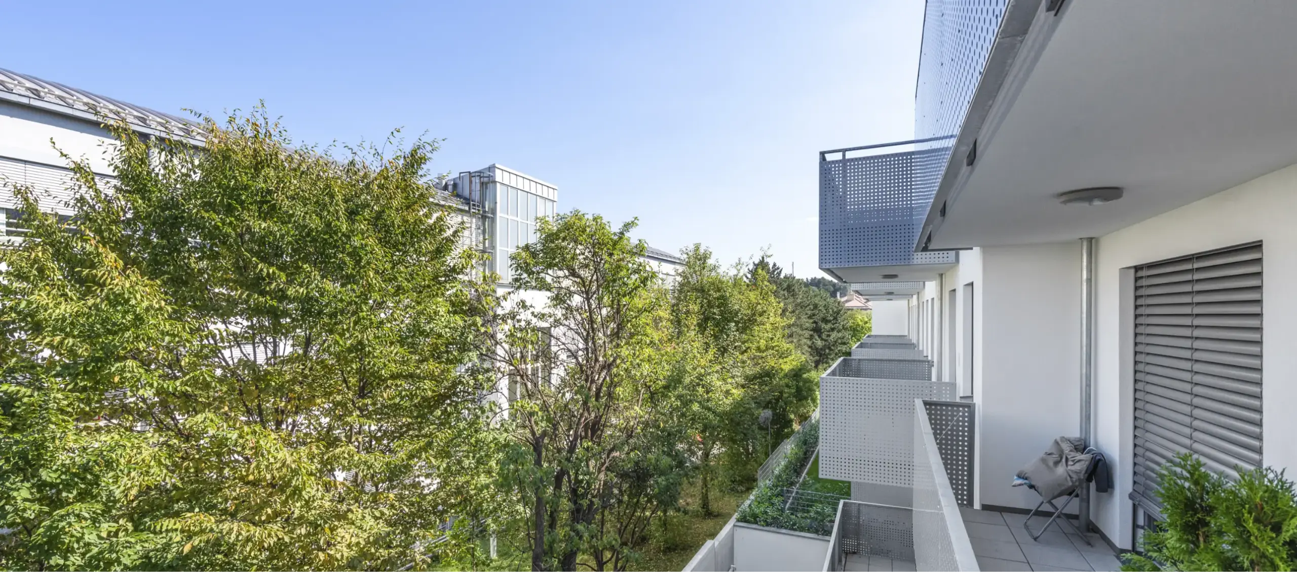 Langer, moderner Balkon mit perforiertem Metallgeländer, umgeben von grünen Bäumen.