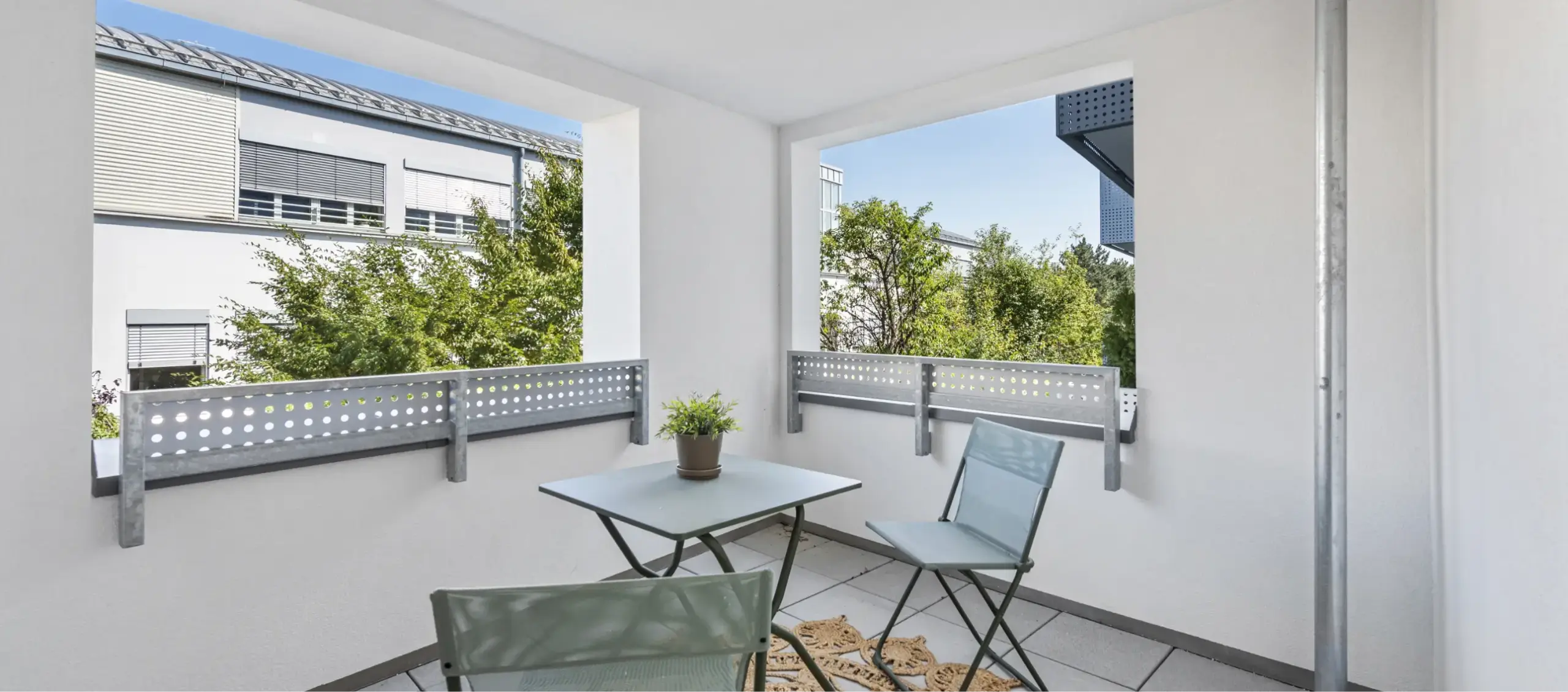 Kleiner Balkon mit Metallmöbeln und grünen Bäumen im Hintergrund.