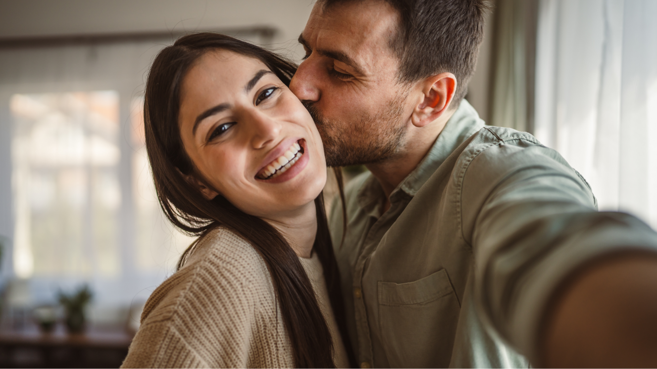 Junges, glückliches Paar macht ein Selfie, Mann küsst Frau auf die Wange.